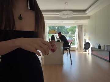 Video of a modern, minimalist living room with light wooden floors, large windows, and a chandelier. A person in a black top and skirt is foreground, while a man in a black shirt sits at a desk, reading.