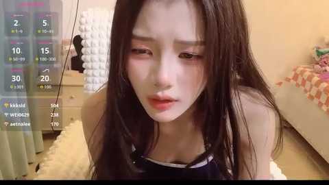 A video of a young Asian woman with long black hair and pale skin, wearing a black top, in a cozy bedroom with a pink and white quilted bedspread and a white nightstand.