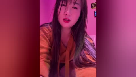 Video of an East Asian woman with long black hair, wearing a mustard-yellow sweater, under a pink lighting. She has a confident expression and red lipstick.