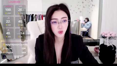 Video of an Asian woman with long black hair, wearing glasses, a black blazer, and sitting at a desk. Background shows a modern office with a mirror and a person in the distance.