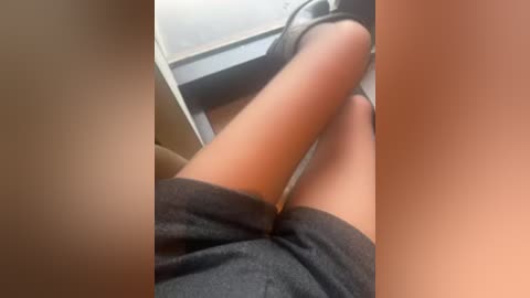 A close-up video shows a person's legs in grey shorts, wearing black sandals, sitting on a beige chair with a white table behind them. The background is blurry.