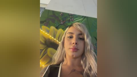 Video of a young woman with long blonde hair, fair skin, and full lips, standing under a large yellow and green floral umbrella. She wears a black top.