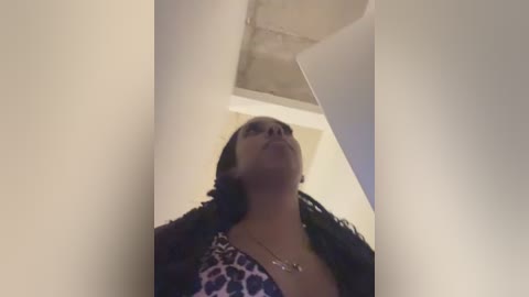 Video of a woman with medium-dark skin, wearing a leopard print top, looking up with a neutral expression. The background is an indoor room with a white ceiling and beige walls.