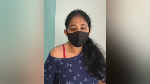 Video of a woman with long black hair, wearing a black mask, blue patterned blouse, and a pink bra strap, standing against a gradient wall.