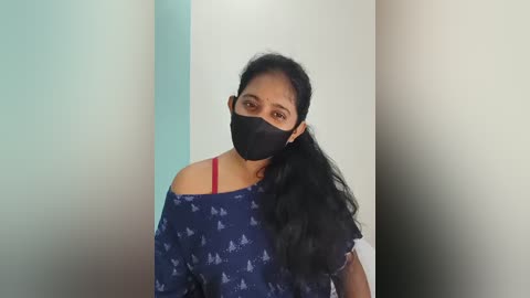 Video of a South Asian woman with medium skin tone and long black hair, wearing a black face mask, red bra strap, and blue patterned top, standing in front of a white and beige wall.
