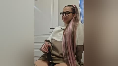 Video of a young woman with long pink dreadlocks, glasses, and a beige sweater, sitting on a white chair in a bright room with white walls and a door.