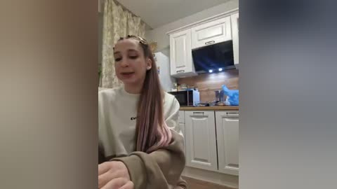 A video of a young woman with long, straight pink hair in a cozy kitchen, wearing a gray sweatshirt, smiling and gesturing towards a partially blurred object in the foreground.
