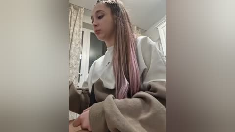 Video of a young woman with long, pastel pink and brown hair, wearing a brown and white sweater, sitting on a couch in a beige room with patterned curtains.