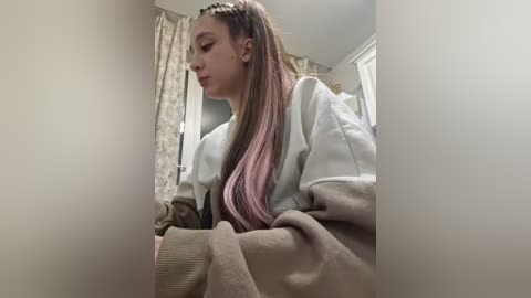Video of a young woman with long, straight, pastel pink hair styled in a braid, wearing a light grey sweater, seated indoors with beige curtains and a window in the background.