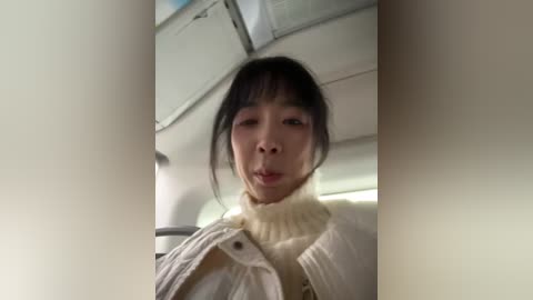 Video of an Asian woman with straight black hair and light skin, wearing a cream-colored turtleneck sweater, smiling, inside an airplane cabin with white walls and overhead bins.
