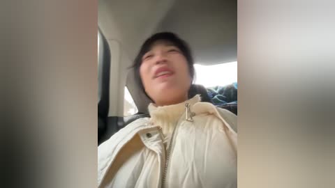 A video of an East Asian woman with short black hair, wearing a beige puffy jacket, smiling, sitting in a car.