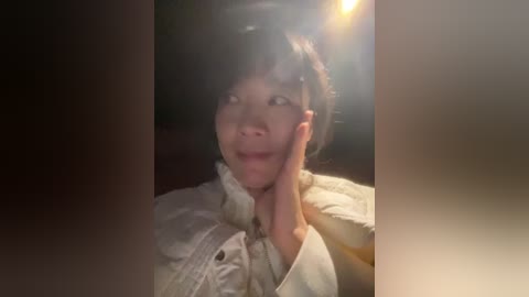 A dimly lit video of an Asian woman with short black hair, wearing a white shirt, resting her chin on her hand in a contemplative pose. The background is blurred and dark.