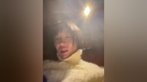 Video of a young, Asian girl with short, dark hair, wearing a fluffy, cream-colored coat, eyes closed, mouth open, surrounded by a warm, blurry, indoor light.