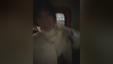A blurry video of a young woman with dark hair and fair skin, smiling, wearing a white turtleneck sweater, indoors, dim lighting.