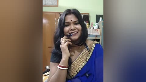 Video of a smiling South Asian woman in a blue and gold sari, wearing a bindi and multiple bangles, holding a phone in a cluttered kitchen with green walls and cabinets.