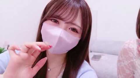 Video of an East Asian woman with long brown hair, wearing a white surgical mask, pink nail polish, and a light blue top, examining a white surgical mask.