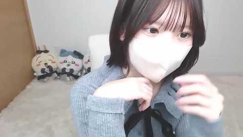 A video of an Asian woman with short black hair, wearing a white mask, a gray sweater, and black bow, adjusting her mask. Background features a white wall, plush toys, and a brown door.