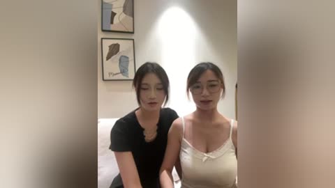 Video of two East Asian women with light skin, one in a black top, the other in a white camisole, sitting side by side in a brightly lit room with modern art on the beige wall.