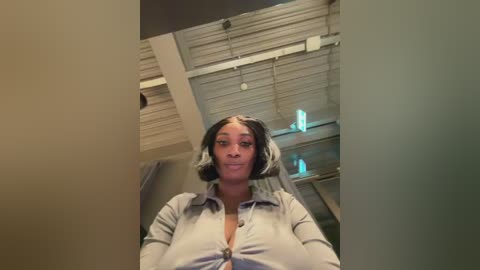 Video of a young Black woman with a medium-dark skin tone, shoulder-length black hair with white streaks, and a grey button-up shirt, standing in a modern, industrial space with exposed ceiling beams and fluorescent lights.