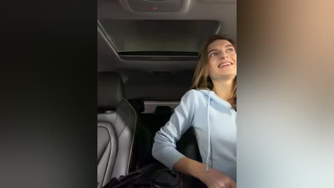 Video of a smiling young woman in a light blue hoodie, seated in a modern car with gray upholstery and tinted windows.