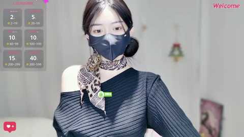 A video of an East Asian woman with fair skin, wearing a black face mask and a leopard-print scarf, in a black ribbed sweater. Background shows a blurred indoor setting with a \"Welcome\" sign.