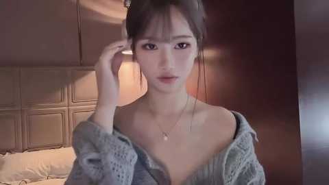 Video of a young Asian woman with fair skin, dark hair in a bun, wearing a loose grey sweater and a delicate necklace, adjusting her hair. She has a serene expression. Background: dimly lit bedroom with a beige, padded headboard and warm, muted tones.