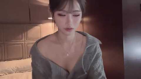 A video of a young East Asian woman with fair skin, small breasts, and medium-sized eyes, wearing a partially unbuttoned green shirt, standing in a dimly lit bedroom with a beige headboard and wooden paneling.