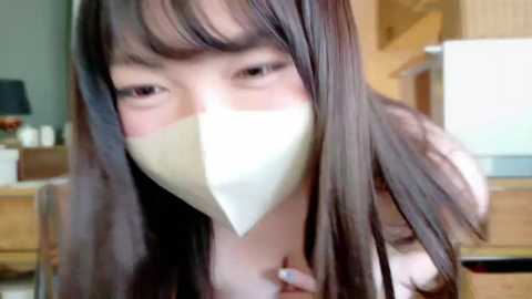 A close-up video of an Asian woman with long black hair, wearing a white surgical mask, and a white shirt, in a modern kitchen setting.