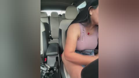 Video of a young woman with long black hair, wearing a lavender sleeveless top and blue jeans, sitting in a car seat, visible from the side.