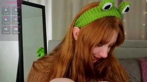 Video of a fair-skinned woman with long, straight, auburn hair wearing a green frog-themed knit headband and a cozy, brown sweater. She is smiling and looking downward. The background includes a white curtain and a mirror.