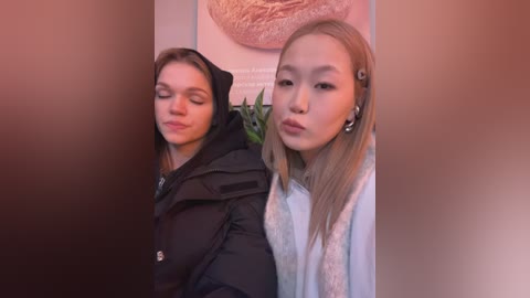 Video of two young Asian women, one with long blonde hair and the other with brown hair, making pouty faces, wearing winter coats, in a cozy indoor setting.