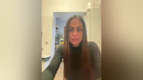 A blurry video of a woman with long brown hair, wearing a black long-sleeve shirt, standing in a dimly lit, cluttered room with shelves and a white wall.