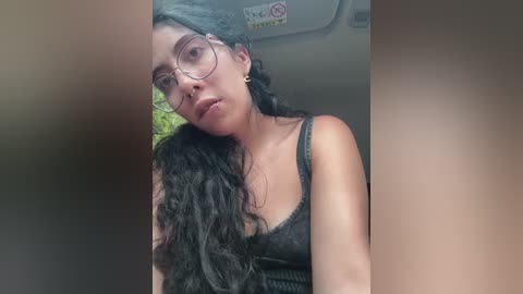 Video of a young woman with long black curly hair, wearing glasses and a dark tank top, leaning against a car window.