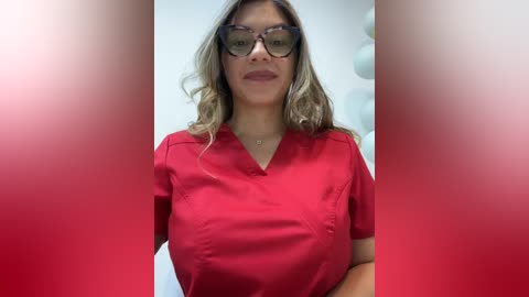 Video of a light-skinned woman with long, wavy blonde hair, wearing black-rimmed glasses, a red scrub top, and a necklace, standing in a clinical setting with a blurred red background.
