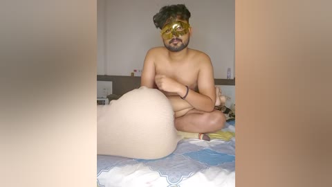 Video of a nude man with light brown skin, wearing yellow goggles, sitting on a bed with a patterned sheet. He has a medium build, short hair, and is covering his groin with his left arm. The room has neutral walls and minimal furniture.