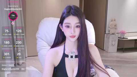Video of an East Asian woman with long black hair, wearing a black halter top, seated in a modern, minimalist room with light wood furniture and a plush chair.