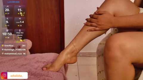 A video captures a woman with light brown skin, sitting nude on a bed with a pink blanket, wearing a gold anklet. The background features a wooden door and a white wall.