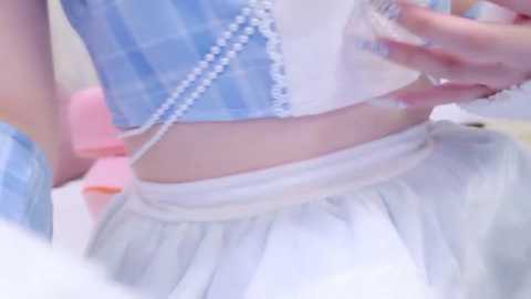 A video of a light-skinned person wearing a blue and white plaid crop top and white skirt, holding a clear plastic bottle with a straw, against a blurred background.
