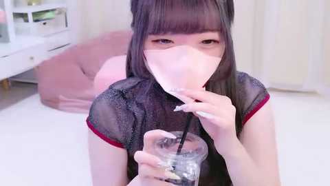 A video of an Asian woman with long, straight black hair, wearing a see-through black top with red trim, drinking from a glass. She is indoors, on a white floor, with a white background.