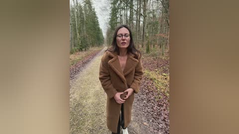 Video of a woman with glasses and long dark hair, wearing a brown, fuzzy coat, standing on a gravel path flanked by tall trees in a forest.