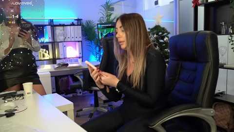Video of a woman with long, straight brown hair in a black outfit, seated at a desk in a modern office, holding a phone, with a colorful computer monitor and shelves in the background.
