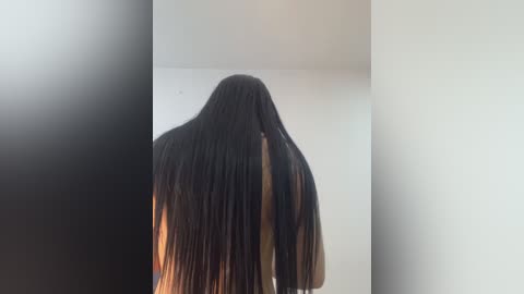 Video of a person with long, straight black hair partially covering their face, standing in a minimalist, light-colored room with plain white walls and a soft, diffused light source.