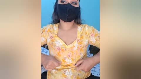 A video of a woman with medium brown skin, wearing a black face mask, floral yellow top, and black pants, sitting against a blue wall, adjusting her shirt.