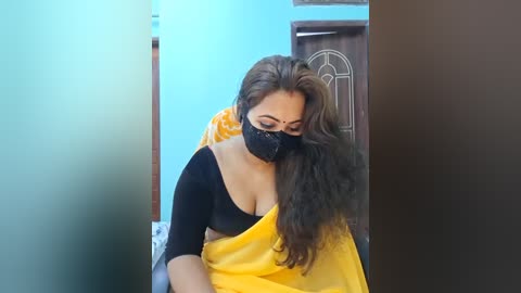Video of a South Asian woman with long brown hair, wearing a black face mask, a black top, and a yellow sari, sitting indoors.