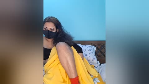 Video of a woman with medium skin tone, wearing a black face mask, black top, and yellow saree, laying on a bed with white and blue patterned pillows, in a room with blue walls.