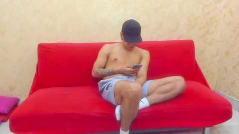 Video of a shirtless, tattooed man with medium skin tone, wearing a black cap and light blue shorts, sitting cross-legged on a bright red couch, absorbed in a smartphone, against a textured beige wall.