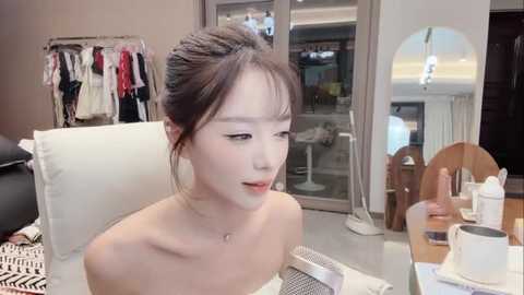 Video of an Asian woman with light skin and long brown hair in a bun, wearing a strapless top, sitting at a table with a cup of coffee and a laptop. Background shows a stylish modern interior with clothes on a rack and a mirror.