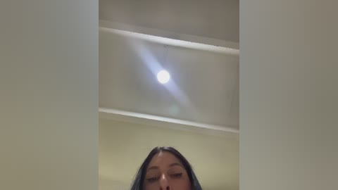 Video of a young woman with long black hair, partially obscured by a vertical slit, focusing on a ceiling light emitting a bright, circular glow.