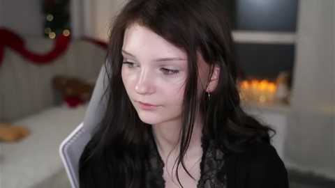 Video of a young woman with long, dark hair, fair skin, and small breasts, wearing a black lace top, standing in a softly lit room with Christmas decorations and candles in the background.