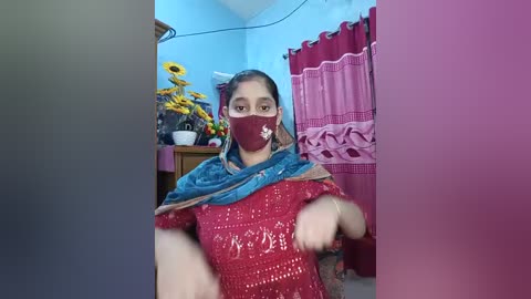 Video of a woman in a red, sequined outfit with a blue shawl, wearing a face mask, sitting on a bed in a room with yellow flowers, pink curtains, and blue walls.